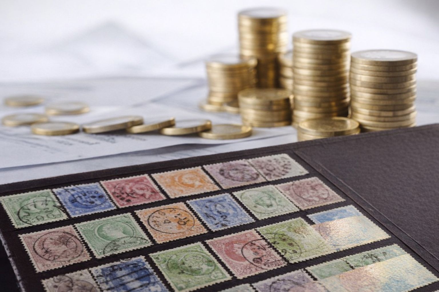 Frimärken i ett album bredvid travar av guldmynt på dokument.